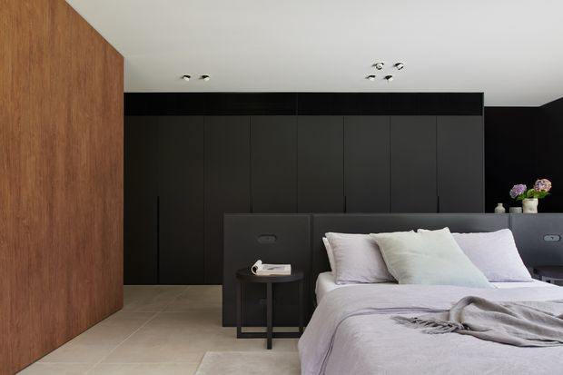 Contemporary bedroom with a low-profile upholstered bed, layered pale bedding, and dark built-in wall cabinetry. A warm timber feature wall contrasts with the matte black cabinetry and dark walls, while recessed ceiling spotlights provide subtle illumination. Neutral tiled flooring and minimal bedside furniture create a restrained, sophisticated interior.