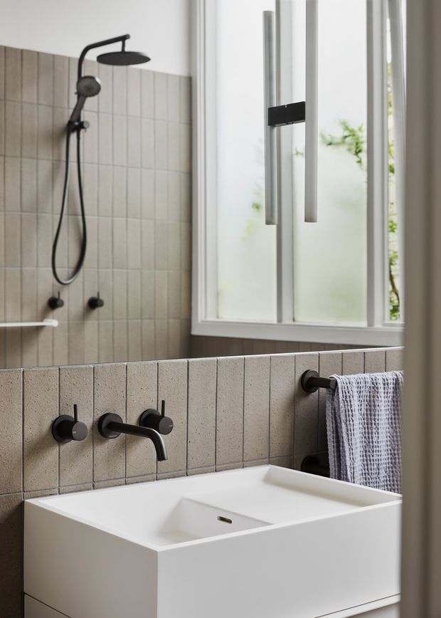 A modern bathroom featuring a wall-mounted white basin with a slim black faucet, beige vertically grooved wall tiles, and matte black shower fittings. A frosted window provides soft natural light, with a white textured towel hanging beside the basin.