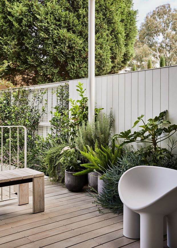 A lush, private outdoor terrace or deck with timber decking, a pale vertical-panel privacy fence, abundant potted greenery, and a simple outdoor dining table with metal chairs. A sculptural white outdoor chair or tub-like seat occupies the foreground, while tall hedging creates a secluded garden atmosphere.