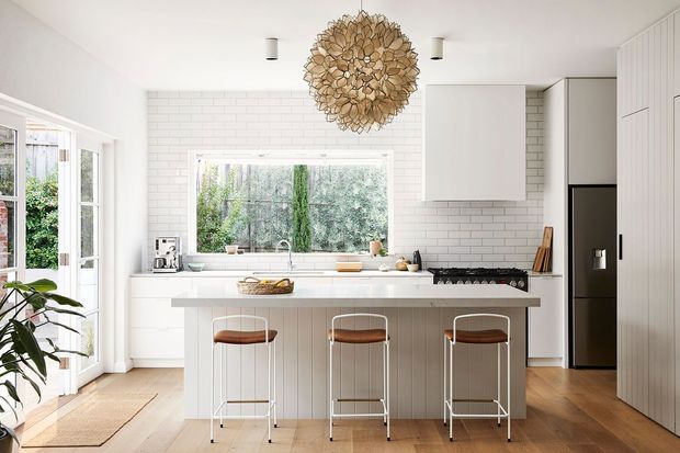 Bright, refined white kitchen with a central island and three bar stools. The space features white subway-tile walls, white shaker-style cabinetry, a white stone benchtop, integrated cooking appliances, a black refrigerator, timber flooring, a large window overlooking greenery, and a sculptural woven pendant light.