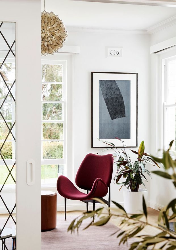 Bright, refined sitting area with white walls, detailed ceiling cornices, tall multi-pane windows, and a full-height glazed or mirrored partition at left. A sculptural burgundy lounge chair sits on a pale rug beside a white planter with leafy greenery, while framed monochrome artwork and a decorative gold pendant add character. The room has a light, elegant, contemporary-traditional feel.