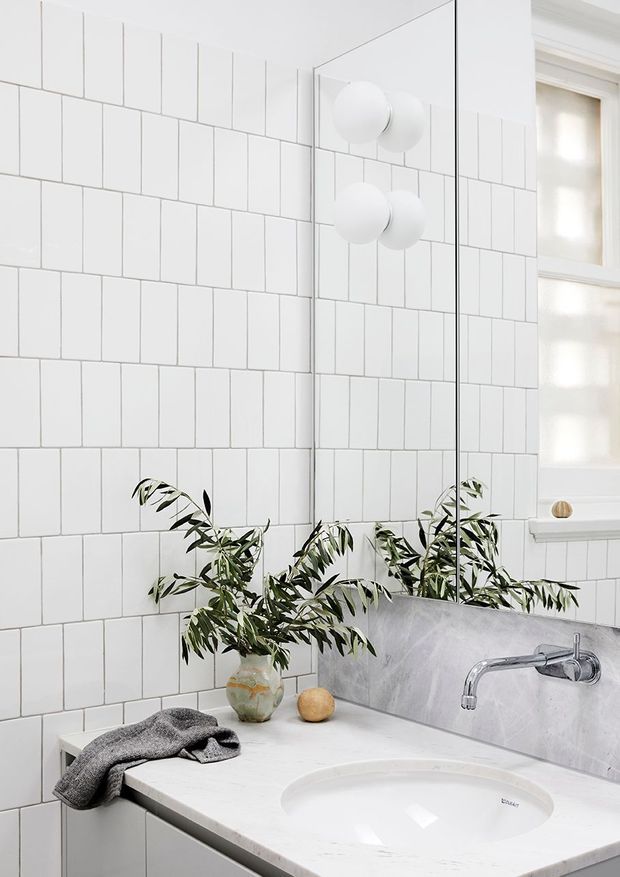 Bright, compact contemporary bathroom featuring white rectangular wall tiles with contrasting grout, a large frameless mirror, spherical wall lights, a white stone vanity benchtop with integrated basin, chrome wall-mounted tapware, and a small frosted window. An olive branch in a ceramic vase adds an organic accent.