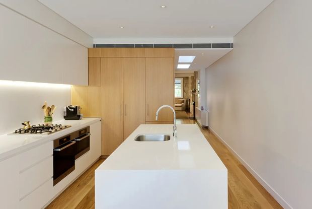A bright, contemporary galley kitchen with a large white island and integrated sink, white cabinetry, warm timber flooring, and full-height timber storage cabinetry. The kitchen includes a gas cooktop, built-in black oven, concealed rangehood, recessed ceiling lights, and skylights. A narrow passage leads toward another interior area at the rear.