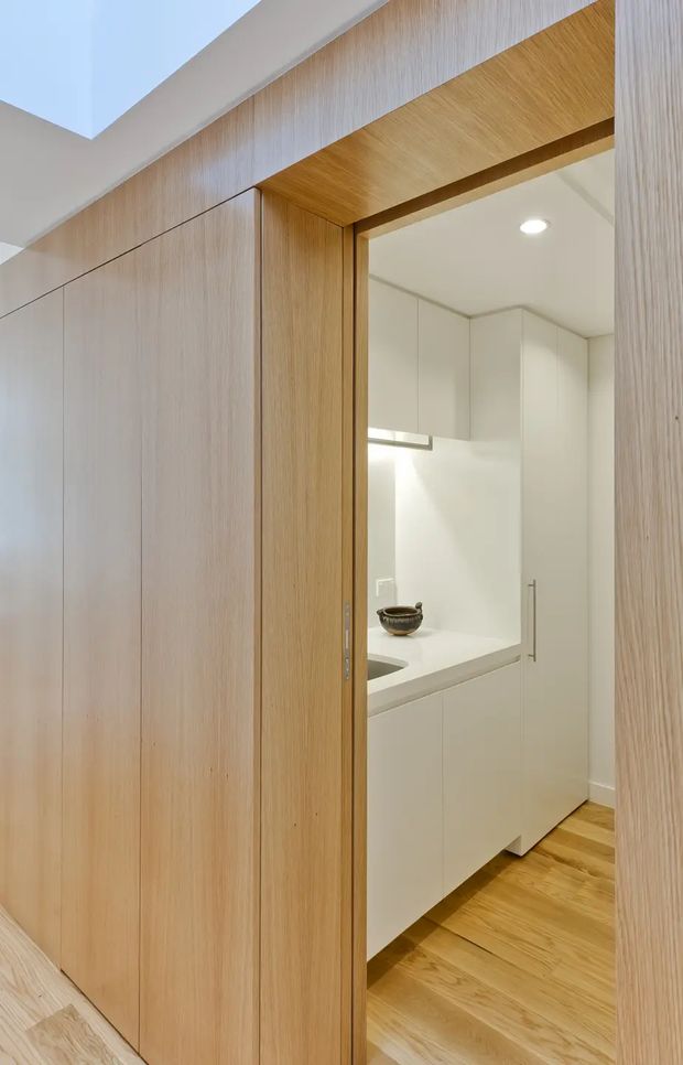 A narrow, minimalist interior view looking through a timber-lined doorway into a compact white kitchen or kitchenette. Full-height light timber wall panelling and concealed cabinetry frame the opening, while the adjoining space contains white handleless cabinets, a pale benchtop, integrated sink, recessed ceiling lighting, and timber flooring.