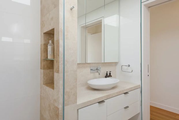 Bright contemporary bathroom featuring a glass-enclosed shower with a built-in tiled niche, a floating white vessel basin on a beige stone-look benchtop, mirrored wall cabinetry, and white vanity drawers. A doorway reveals timber flooring and an adjacent interior space.