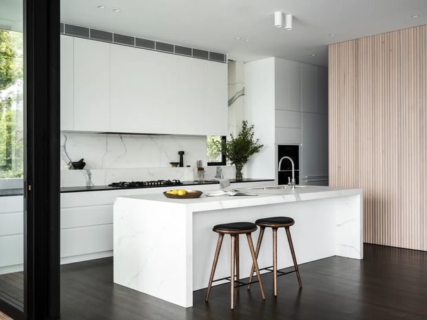 Bright contemporary kitchen with a large white waterfall island, integrated white cabinetry, marble-look splashback, dark timber flooring, black-framed glazing, and a vertical timber slat feature wall. Two timber bar stools sit at the island, with recessed ceiling lighting and integrated appliances creating a clean, refined interior.