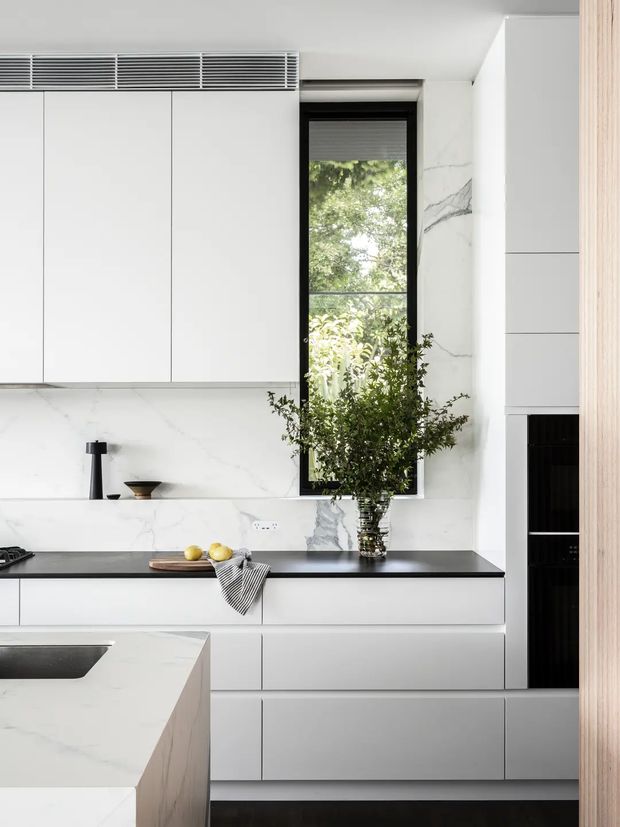 A bright, refined contemporary kitchen with extensive white flat-panel cabinetry, veined white stone wall surfaces and benchtops, a dark contrasting rear benchtop, and a central black-framed window overlooking greenery. A large island with an inset sink occupies the foreground, while integrated ventilation and tall storage create a streamlined built-in composition. Natural greenery and small accessories add warmth to the otherwise monochromatic space.
