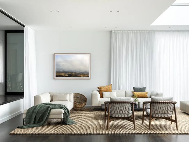 Bright, airy contemporary living room with white walls and full-height sheer curtains, furnished with two sofas, woven lounge chairs, a textured area rug, and a low coffee table. A skylight and recessed ceiling lights provide additional daylight, while a framed landscape artwork and warm mustard cushions add subtle colour. Dark timber flooring and black-framed glazing are visible at the edges.
