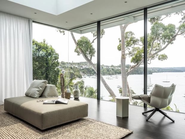 Bright, refined living room with floor-to-ceiling corner glazing overlooking a leafy waterfront and boats. The room features a large neutral upholstered ottoman, sculptural lounge chair, sheer white curtains, dark timber flooring, a textured natural-fibre rug, and a minimalist recessed ceiling with skylight-like opening.