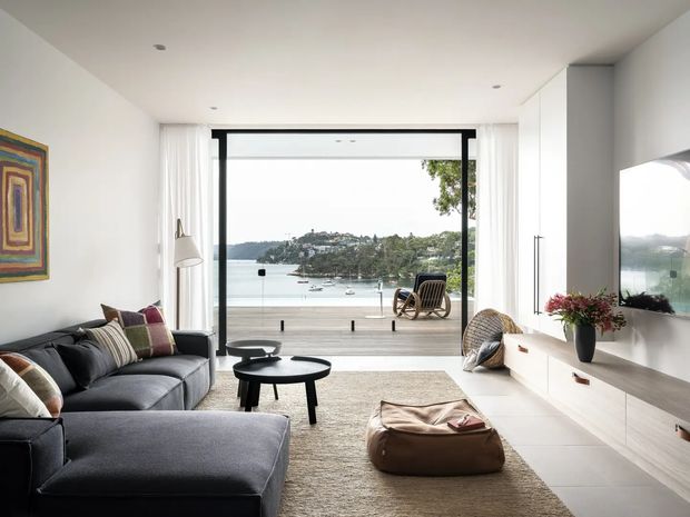 Bright contemporary living room opening onto a waterfront terrace, with full-height black-framed glazing, white walls and ceiling, a dark charcoal sectional sofa, round coffee tables, a large neutral rug, timber-toned built-in media cabinetry, and a wall-mounted television. The view shows a calm bay and wooded shoreline beyond.