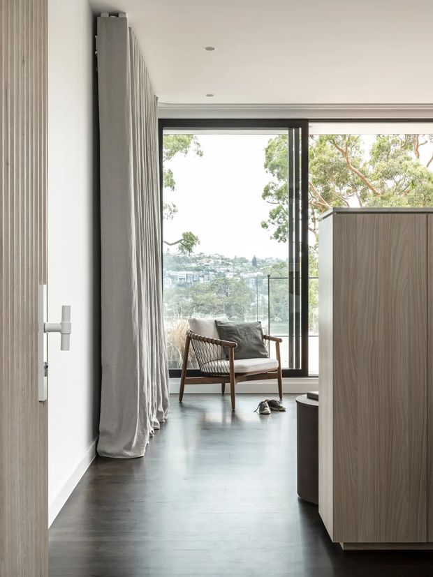 A calm, minimalist interior view looking through a doorway toward a bright sitting area. The space features dark timber flooring, white walls and ceiling, full-height grey curtains, black-framed glazing, and a timber built-in cabinet. A mid-century style lounge chair sits by the window overlooking leafy suburban scenery.