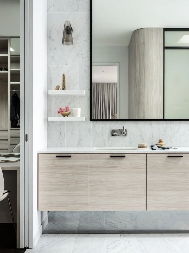 A refined contemporary bathroom vanity with a floating light timber-grain cabinet, white marble-look wall and floor tiles, integrated white benchtop, black-framed mirrored storage, chrome wall-mounted tapware, and open display shelves. A partially visible adjoining room with wardrobe joinery and a chair appears through the doorway.