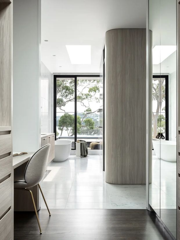 A bright, refined bathroom viewed from an adjoining dressing or vanity area. The space features a freestanding white bathtub positioned in front of expansive black-framed windows, a large mirrored wall, pale stone or porcelain flooring, white walls, and a prominent curved vertical timber-look partition or built-in element. A small vanity desk with an upholstered chair is visible in the foreground, while a second bathtub or bathroom zone appears in reflection. The room has abundant natural light and an open, spa-like feel.