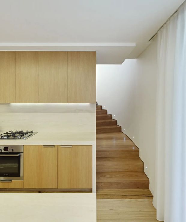 A bright, minimalist open-plan kitchen with pale timber cabinetry, a light stone or solid-surface splashback and benchtop, and an integrated gas cooktop and oven. Beside the kitchen, a warm timber staircase rises along a white wall, with small recessed step lights and full-height sheer curtains at the right. The space has clean lines, concealed storage, and a restrained natural-material palette.