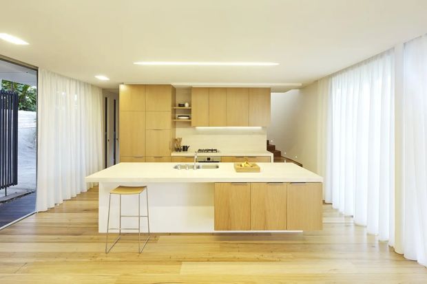 Bright, minimalist open-plan kitchen with a large white island, integrated sink and cooktop, pale timber cabinetry, and extensive sheer white curtains along full-height glazing. A timber staircase is visible behind the kitchen, with a small exterior glimpse through the left opening.