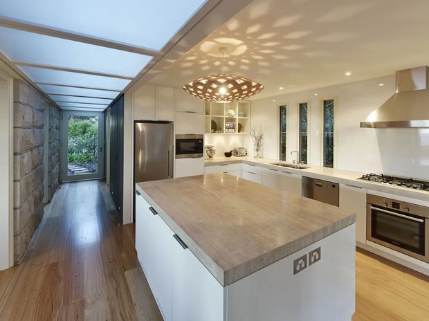 Bright contemporary kitchen with a large central island, white flat-panel cabinetry, timber-look benchtop, stainless-steel appliances, and a sculptural perforated pendant light. A glazed corridor with a stone feature wall and timber flooring extends alongside the kitchen, with garden greenery visible at the far end.