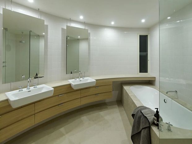 A spacious contemporary bathroom featuring a long timber-toned floating vanity with two white rectangular basins, large mirrored panels, pale tiled walls and floor, recessed ceiling lights, and a built-in bathtub enclosed by a glass shower screen. A narrow vertical window provides additional natural light.