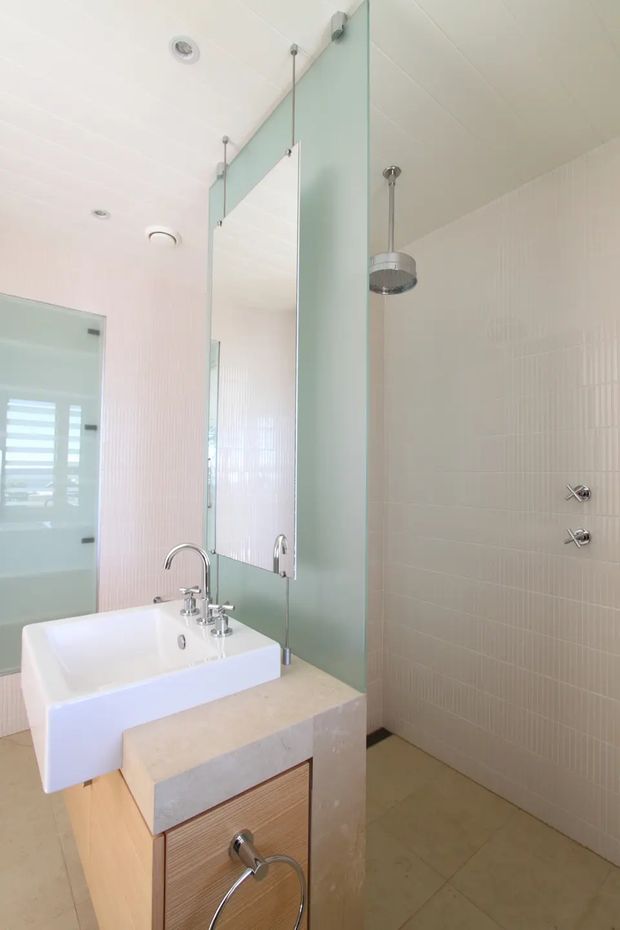 Bright contemporary bathroom featuring a white vessel basin on a pale stone vanity, timber-toned drawer cabinetry, a large frameless mirror, glass shower enclosure, chrome fixtures, and light vertically textured wall tiles. The space has a clean, airy appearance with recessed ceiling lighting and a suspended showerhead.