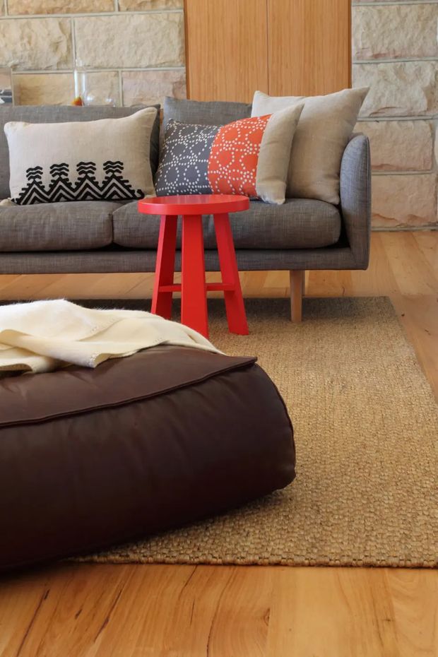 A warm, intimate living room vignette featuring a grey upholstered sofa with patterned neutral and red cushions, a bright red side table, and a large dark brown leather ottoman. The room has honey-toned timber flooring, a natural woven rug, a light stone feature wall, and vertical timber wall panelling. The styling is eclectic and mid-century-influenced with strong red accents.