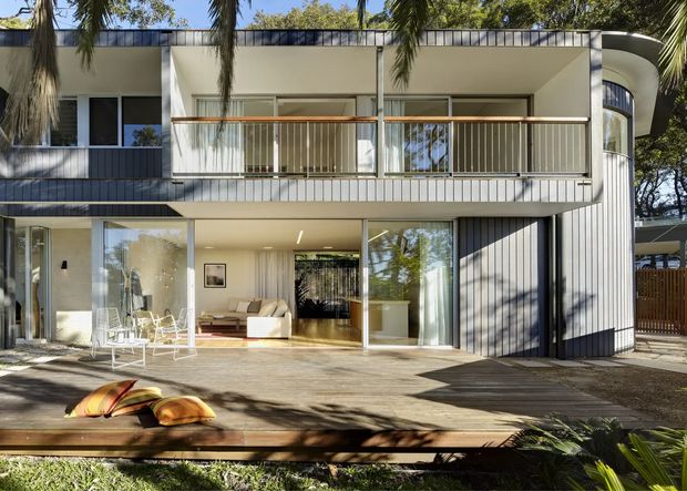 Exterior view of a contemporary multi-level residence opening onto a broad timber deck. The facade combines pale vertical cladding, extensive glazing, white rendered surfaces, dark balcony panels, and slim metal balcony railings. A curved glazed volume is visible on the right, while the ground level opens to a light-filled living area with furniture visible inside. Mature palms and trees surround the deck, creating a leafy setting.