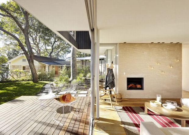 Open-plan living area with floor-to-ceiling sliding glazing opening onto a timber deck and landscaped garden. The interior features a pale stone fireplace wall with inset firebox, timber flooring, a low coffee table, lounge seating, and a striped rug. The deck has lightweight outdoor dining furniture and connects visually to a low single-storey house surrounded by mature trees.
