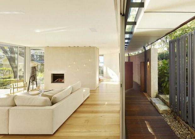 Bright modern open-plan living room with a large cream sectional sofa, pale timber flooring, extensive glazing, and a minimalist marble-like fireplace surround. A covered outdoor walkway with timber decking, vertical screening, stone edging, and planted greenery runs alongside the glazed interior.