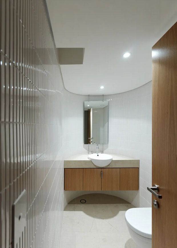 Compact contemporary bathroom viewed through a partially open timber door. The room features white vertically textured wall tiles, a curved white ceiling with recessed downlights, pale tiled flooring, a floating timber vanity with a light stone benchtop, a round vessel basin, wall mirror, and a partially visible toilet.