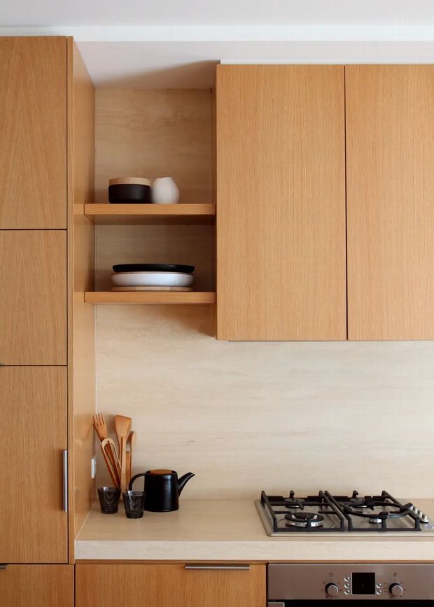 A close-up view of a refined modern kitchen with warm timber cabinetry, a pale stone or composite benchtop and matching light backsplash. Full-height cabinetry frames open timber display shelves holding ceramics, while a gas cooktop and stainless-steel built-in oven occupy the lower cabinetry. Cooking utensils, a black kettle and small cups sit on the benchtop. The composition is clean, warm and highly cohesive, with simple flat-front joinery and minimal detailing.