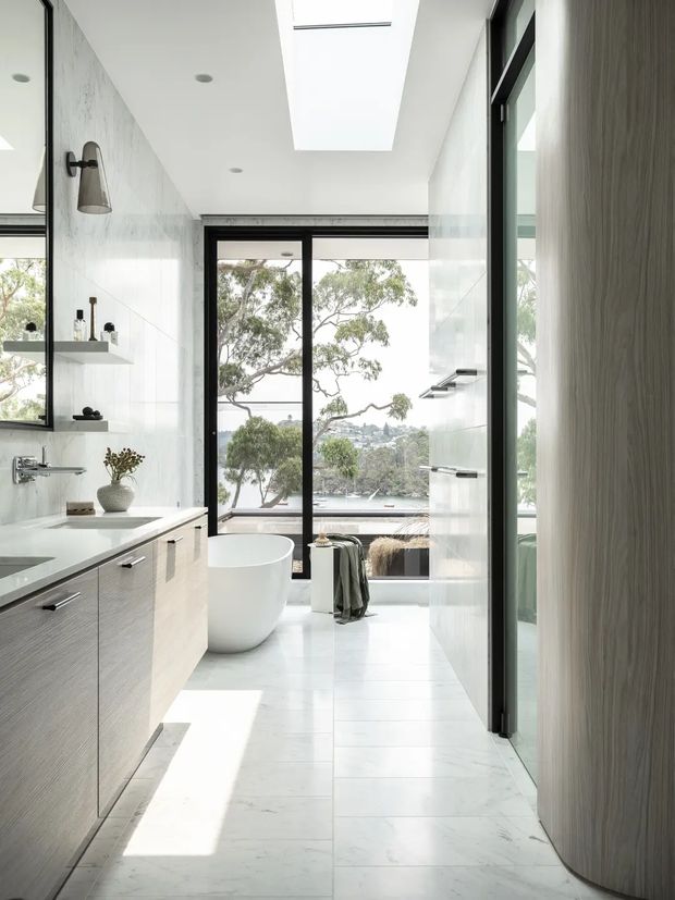 Bright contemporary bathroom with a freestanding white bathtub positioned beside full-height black-framed glazing overlooking a leafy waterfront setting. The room features pale marble-look wall and floor tiles, a floating light-wood vanity with integrated white benchtop, black-framed glass shower enclosure, wall-mounted fixtures, a large skylight, and a curved wood-toned door or wall panel.