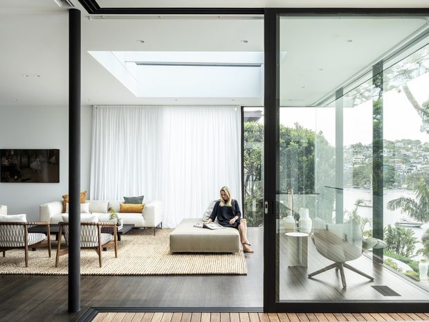 Bright contemporary living room viewed through large black-framed glazed sliding doors, with a skylight, sheer curtains, cream sofas, woven rug, upholstered ottoman, and a separate glass-enclosed seating or workspace area. Dark timber flooring and a small timber deck threshold contrast with the light interior. The glazing offers views toward a leafy urban setting.