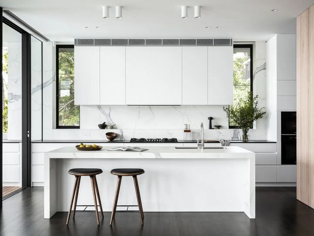 Bright contemporary kitchen with a large white waterfall-edge island, two timber-and-black bar stools, white flat-panel cabinetry, marble-look splashback, integrated appliances, and dark flooring. Black-framed glazing and a timber wall panel add contrast, while concealed and recessed lighting keep the space clean and minimal.