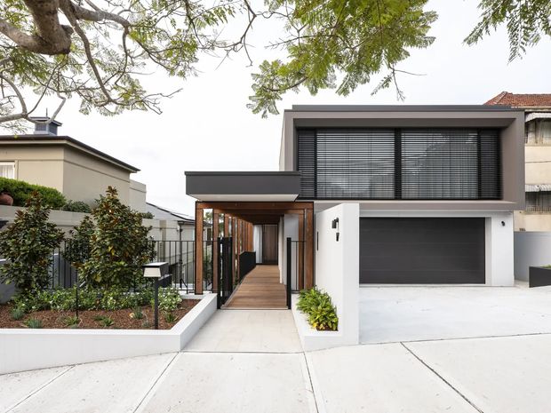 Front exterior view of a contemporary two-storey residence with a flat-roofed form, dark rendered upper facade, broad horizontal louvres over the upper glazing, and an integrated double garage. A timber-lined recessed entry walkway leads from the landscaped front garden to the entrance, with pale concrete paving and low white boundary walls.