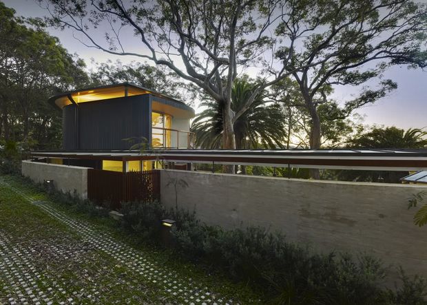 Exterior view of a contemporary multi-level residence nestled among mature trees. The building features dark vertical cladding, expansive glazed openings, a projecting flat roof with warm concealed lighting, and a long low concrete boundary wall. A permeable paved driveway and landscaped planting occupy the foreground, while a large palm and established canopy trees surround the home.