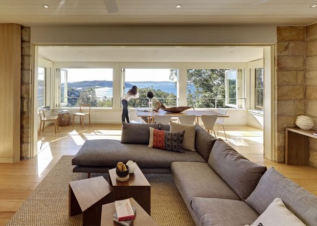 A bright, open-plan living and dining area with a large grey sectional sofa in the foreground, timber coffee tables, a textured rug, and a dining table positioned beside expansive windows. The room opens to a panoramic water and landscape view, with warm timber flooring, pale timber ceilings, stone-clad side walls, and extensive glazed doors and windows. The interior has a relaxed, refined contemporary character with strong indoor-outdoor connection.