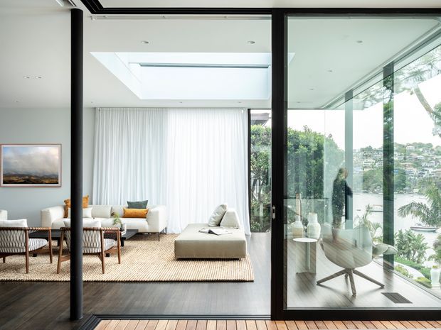 A bright, contemporary living room viewed through full-height black-framed glazing and sliding doors. The room contains white sofas, a large upholstered ottoman, woven lounge chairs, a neutral textured rug, sheer curtains, and dark timber flooring. An adjacent glazed home office with a desk and task chair is visible to the right, while greenery and neighbouring homes can be seen outside. A large overhead skylight or glazed ceiling opening brings in abundant natural light.