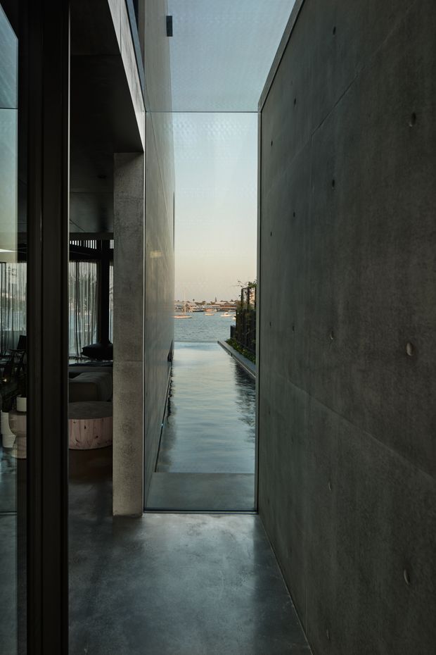A narrow, minimalist concrete passage beside a glazed interior space, terminating in a water view. The corridor features dark polished concrete flooring, smooth grey concrete walls with exposed circular form-tie marks, full-height glazing, and a shallow linear water feature or pool extending toward the waterfront. A dark-toned living area is partially visible through the glazing on the left.