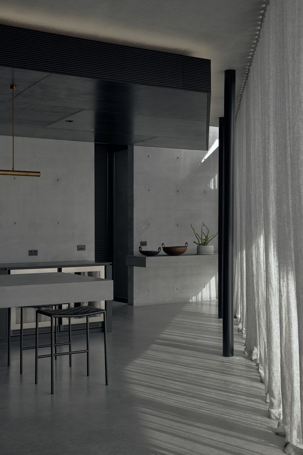 A moody minimalist interior showing a concrete kitchen and dining area. The space features a large concrete island with dark stools, a dark linear ceiling vent or louvre, black structural and joinery elements, a slim pendant light, a concrete feature wall with a shallow shelf and decorative vessels, and full-height sheer curtains casting strong linear shadows across the polished concrete floor.