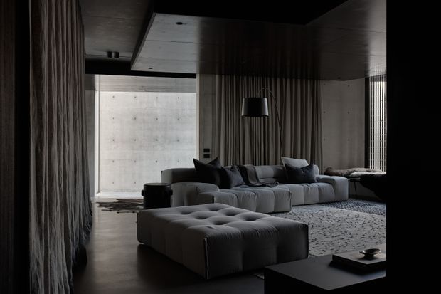 A moody, sophisticated contemporary living room with a large low-profile sectional sofa and tufted ottoman arranged around a dark coffee table. Full-height drapery, dark ceiling and walls, concrete surfaces, soft indirect light, and patterned carpeting create a dramatic, refined atmosphere. A bright concrete courtyard or exterior wall is visible through the glazing.