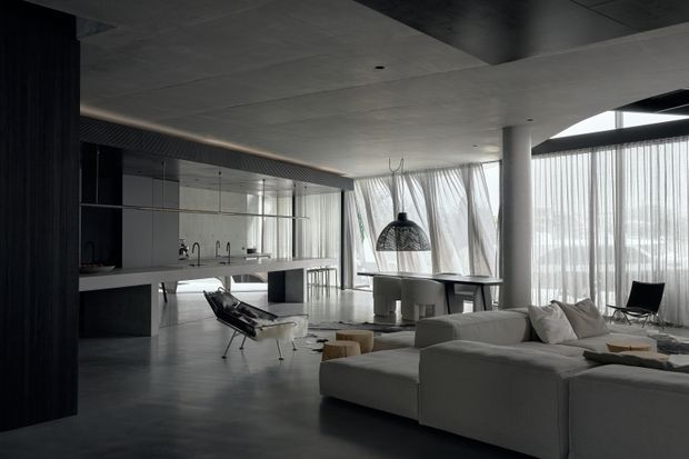 A spacious open-plan contemporary interior combining a kitchen, dining area and living room. The room features a dark minimalist kitchen with a long island, pale sectional sofa, concrete-look ceiling and floor, full-height sheer curtains, slim structural columns, and a dramatic woven pendant light. The muted monochrome palette and abundant glazing create a refined, moody atmosphere.