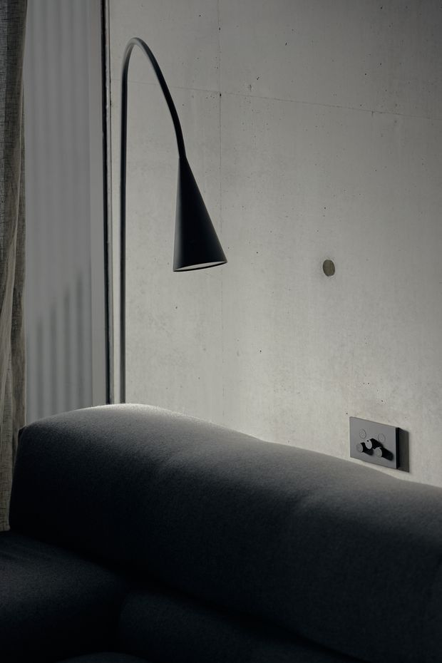 Close-up interior detail of a dark charcoal upholstered sofa against a pale exposed concrete wall. A slim black sculptural wall lamp, dark electrical outlet, and narrow window or curtain edge are visible. The composition is minimalist, moody, and focused on refined lighting and material contrast.