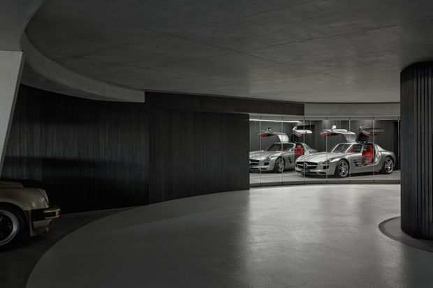 A dramatic, dimly lit indoor automotive showroom or private garage with a broad polished grey concrete floor, curved concrete ceiling, and dark vertical timber or ribbed wall cladding. Several silver sports cars are displayed behind full-height glazing or mirrors at the rear, while part of a vintage vehicle is visible at left. The space has a sculptural, curved circulation layout and a high-contrast industrial-modern atmosphere.