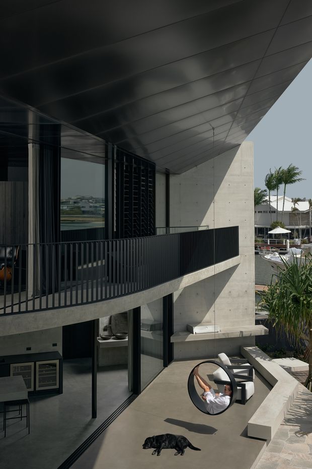 A contemporary multi-level concrete residence viewed from an elevated outdoor terrace. The building features a dramatic cantilevered dark metal roof, exposed concrete walls, extensive black-framed glazing, and a black vertical-bar balcony balustrade. Below, a paved outdoor lounge area includes a circular hanging chair, outdoor seating, a long concrete bench, and integrated planting. A pool or resort-like outdoor setting is visible in the background.