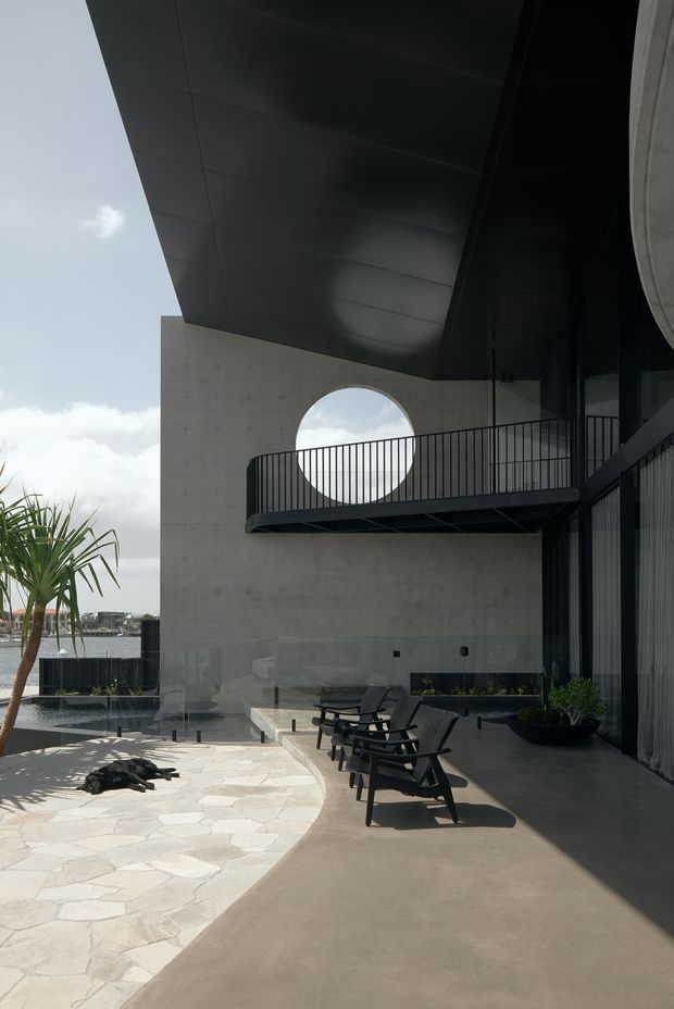 A dramatic modern outdoor terrace or courtyard beside a body of water, featuring a large cantilevered dark metal roof, raw concrete walls, full-height dark glazing, a black metal balcony railing, and a distinctive circular opening in the concrete wall. The terrace has lounge chairs, sculptural black planters, pale irregular stone paving, and a minimalist pool or water feature. The composition emphasizes strong geometric forms, deep overhangs, and contrasting light and shadow.
