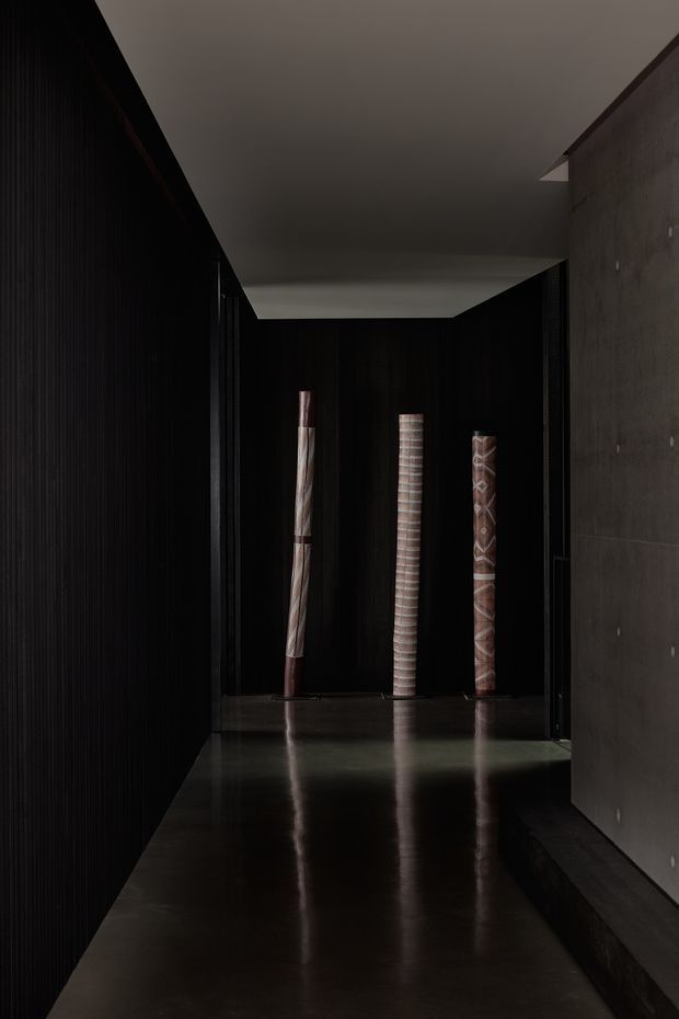A dark, minimalist interior corridor or gallery-like passage with a highly reflective dark floor. The left wall is covered in black vertical ribbed panelling, while the right wall is a raw grey concrete surface. Three tall patterned cylindrical artworks or decorative objects stand against a black feature wall at the end of the passage. The space is dramatically lit with subdued, moody illumination and strong reflections.