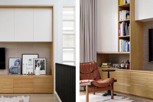 A refined contemporary living room with built-in timber joinery spanning the walls, including open shelving, concealed cabinetry, and horizontal slatted storage. A brown leather lounge chair, framed artwork, books, a television, and soft full-height curtains add warmth. A narrow vertical window and dark radiator are visible between the joinery elements.