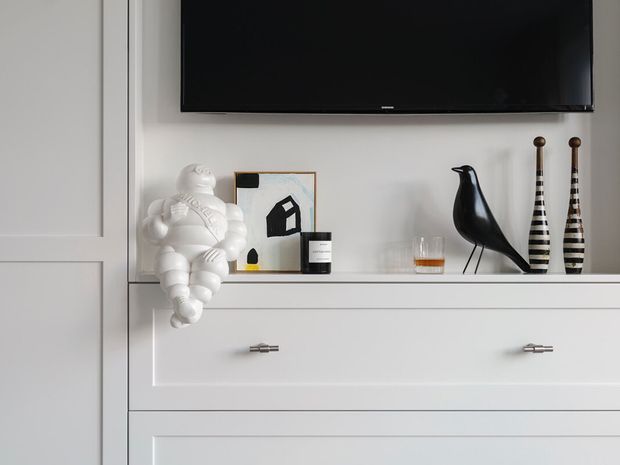 Close-up of a refined white built-in media cabinet and wall paneling beneath a wall-mounted black television. The cabinetry has recessed panel fronts and slim metal pulls, with a curated arrangement of sculptural and graphic decorative objects on the open shelf, including a black bird figure, striped vessels, artwork, and a small drink glass.
