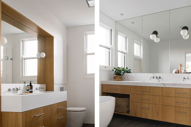 Bright contemporary bathroom with a floating timber vanity, white integrated basin and stone countertop, large wall mirror, white tiled or painted walls, black floor tiles, twin windows, and spherical wall lighting. A freestanding toilet is visible beside the vanity.