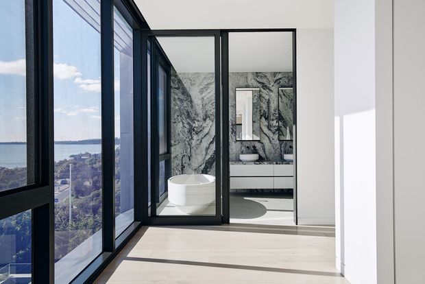 Bright contemporary interior with floor-to-ceiling black-framed glazing overlooking a coastal or waterside urban landscape. A compact bathroom is visible through black-framed glass doors, featuring dramatic grey-and-white marble-look wall surfaces, a freestanding white bathtub, wall-mounted vanity, and large mirror. Pale timber flooring and crisp white walls create a light, refined atmosphere.