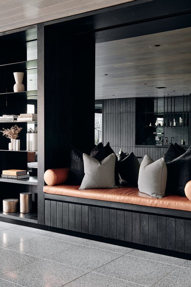 A refined dark-toned interior featuring a built-in black timber banquette with a caramel leather seat, patterned and black cushions, and integrated open shelving displaying books and decorative objects. Large grey stone or terrazzo-look floor tiles, dark vertical timber wall panelling, exposed timber ceiling boards, and a mirrored or glazed wall create a sophisticated lounge or entry setting.