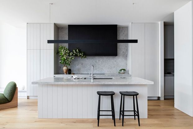 Bright contemporary kitchen with a central island clad in vertical white panels, a pale stone benchtop, and two black bar stools. The rear wall features integrated white cabinetry, a grey textured stone backsplash, a black rangehood and dark cabinetry, with timber flooring and a partially visible green lounge chair.